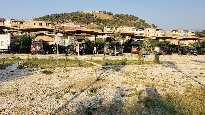 Grassy pitches for caravans, motorhomes, and tents at Riverside Camping Berat, surrounded by greenery with Berat Castle visible on the hill in the background.