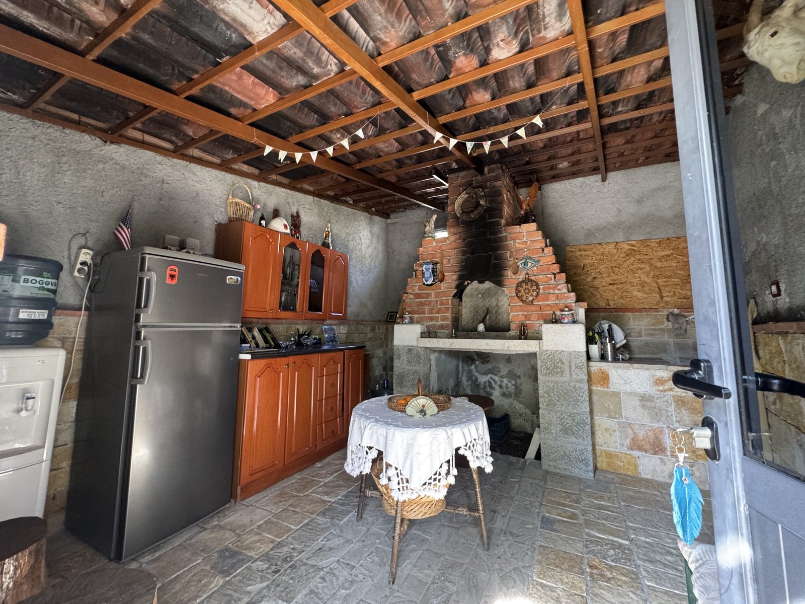 Rustic shared kitchen at Riverside Camping Berat featuring a central stone fireplace, drinking water tap, fridge, mountain stone tile sink, kitchen cabinets, and a traditional Albanian coffee table.